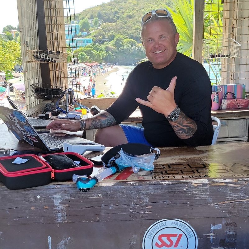 Man at dive center desk with laptop, gives shaka gesture, beach in background.